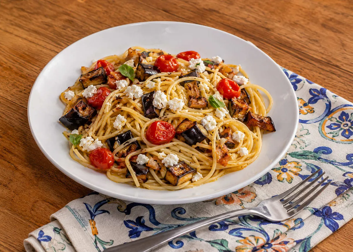 Espaguetis con berenjena asada, tomates cherry y feta desmenuzado en un plato blanco sobre una servilleta estampada